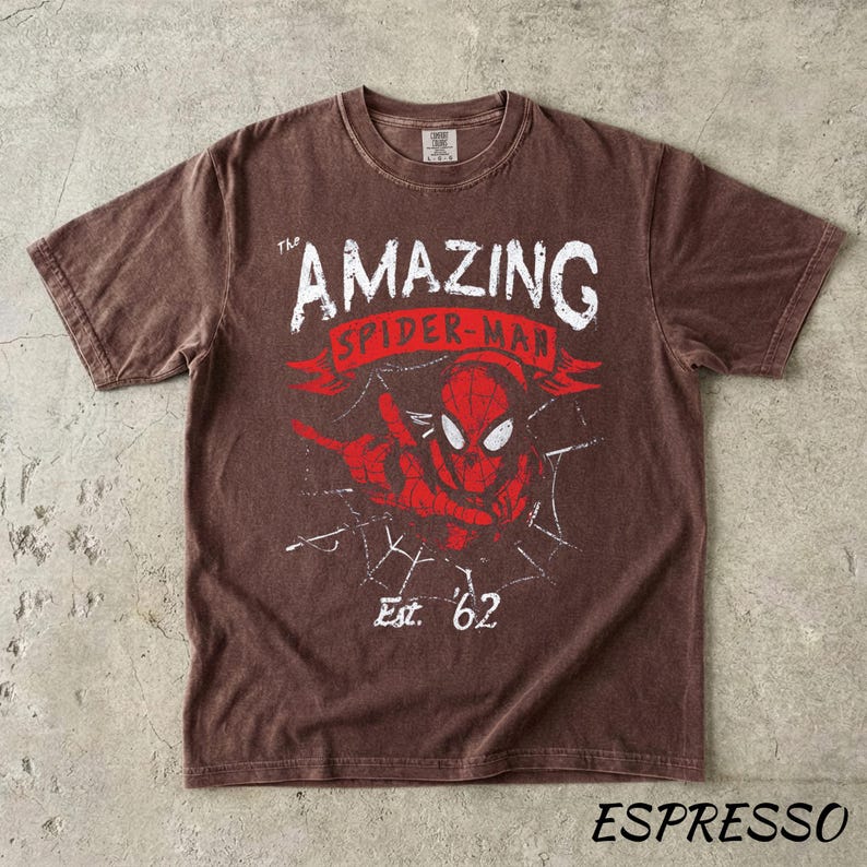 Puede incluir: Camiseta color espresso con un gr&aacute;fico desgastado de Spider-Man. El dise&ntilde;o incluye el texto "THE AMAZING SPIDER-MAN" en blanco y rojo, una telara&ntilde;a y "Est. '62".
