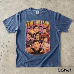 May include: Denim blue t-shirt featuring a graphic of Tom Holland in various poses, with the text "TOM HOLLAND" in a retro style. The shirt has a crew neck and short sleeves. The word "DENIM" is printed at the bottom.