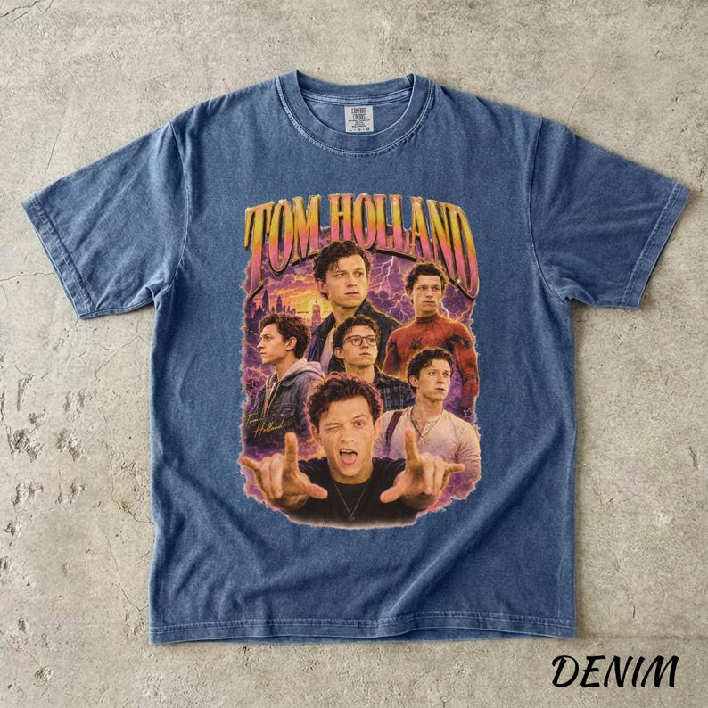 May include: Denim blue t-shirt featuring a graphic of Tom Holland in various poses, with the text "TOM HOLLAND" in a retro style. The shirt has a crew neck and short sleeves. The word "DENIM" is printed at the bottom.