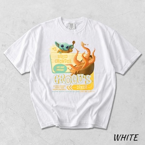 Puede incluir: Camiseta blanca con un dise&ntilde;o de dibujos animados de Grogu's Diner, que sirve sopa de calamares. El dise&ntilde;o incluye un personaje de dibujos animados, tent&aacute;culos naranjas y el texto "Squid Chowder Life Day Special!"