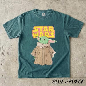 May include: Teal-colored t-shirt featuring a graphic of Grogu from Star Wars, with the Star Wars logo in pink and yellow. The text "BLUE SPURCE" is printed at the bottom of the shirt.