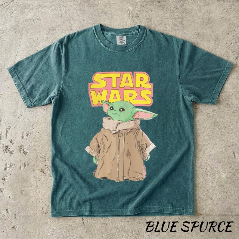 May include: Teal-colored t-shirt featuring a graphic of Grogu from Star Wars, with the Star Wars logo in pink and yellow. The text "BLUE SPURCE" is printed at the bottom of the shirt.