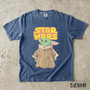 May include: Denim blue t-shirt featuring a graphic of Grogu from Star Wars. The design includes the Star Wars logo in pink and yellow. The image of Grogu is in green, tan, and white. The word "DENIM" is printed at the bottom.
