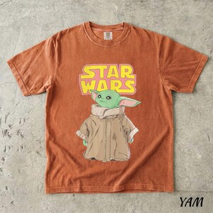 May include: Burnt orange t-shirt featuring a graphic of a green alien character from Star Wars, with the text "STAR WARS" in yellow and pink. The character is wearing a brown coat. The shirt has a crew neck and short sleeves.