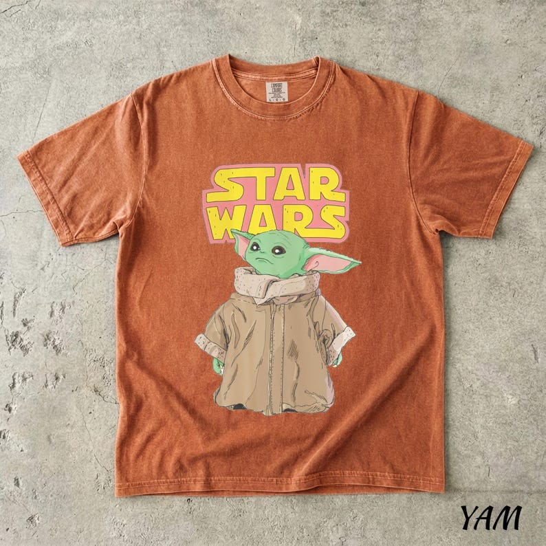 May include: Burnt orange t-shirt featuring a graphic of a green alien character from Star Wars, with the text "STAR WARS" in yellow and pink. The character is wearing a brown coat. The shirt has a crew neck and short sleeves.