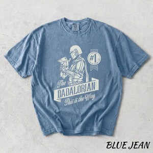 Pu&ograve; includere: Una maglietta blu jeans con una grafica di un personaggio di una popolare serie di fantascienza. La grafica include il testo "The DADALORIAN This is the Way" e un'immagine di un personaggio che tiene una piccola creatura. La maglietta ha un girocollo.