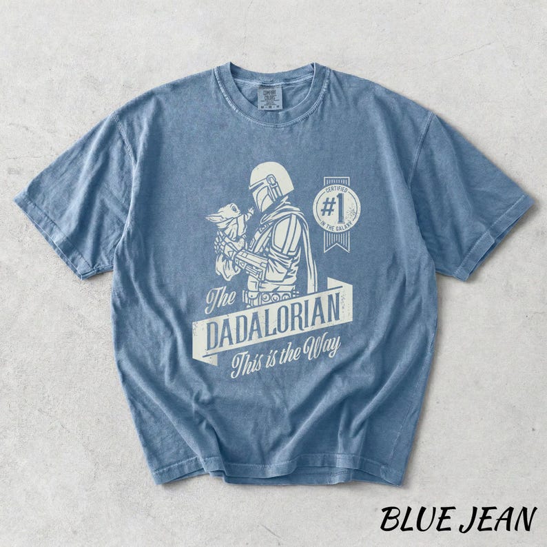 Pu&ograve; includere: Una maglietta blu jeans con una grafica di un personaggio di una popolare serie di fantascienza. La grafica include il testo "The DADALORIAN This is the Way" e un'immagine di un personaggio che tiene una piccola creatura. La maglietta ha un girocollo.
