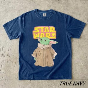 May include: Navy blue t-shirt featuring a graphic of a green alien character from Star Wars, with the text "STAR WARS" in yellow and pink. The shirt is labeled "TRUE NAVY" at the bottom.