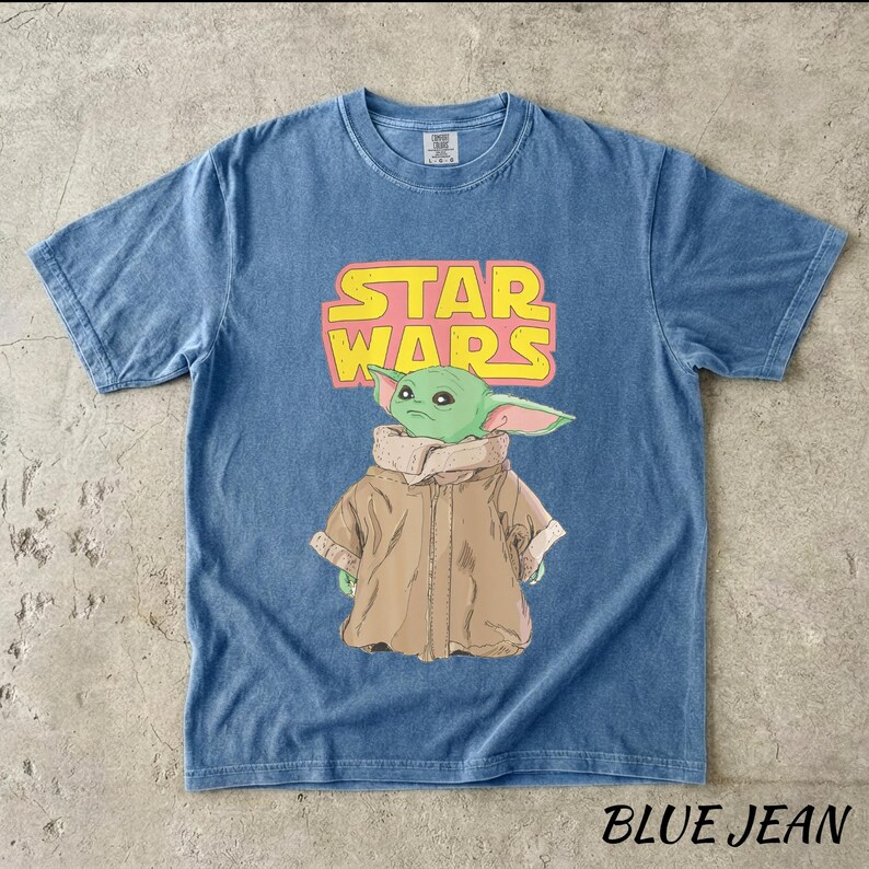 May include: Blue jean t-shirt featuring a graphic of the Star Wars logo and a green alien character. The graphic is in yellow and pink. The text "BLUE JEAN" is at the bottom of the shirt.