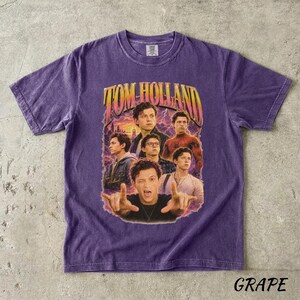 May include: A purple t-shirt featuring a graphic of Tom Holland with the text "TOM HOLLAND" in a retro style. The design includes multiple images of the actor. The word "GRAPE" is printed at the bottom of the shirt.