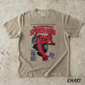 May include: Khaki-colored t-shirt featuring a vintage comic book graphic of Spider-Man. The design includes the text "The Amazing Spider-Man" in red and black, along with comic book panels. The shirt is a crew neck and has short sleeves.