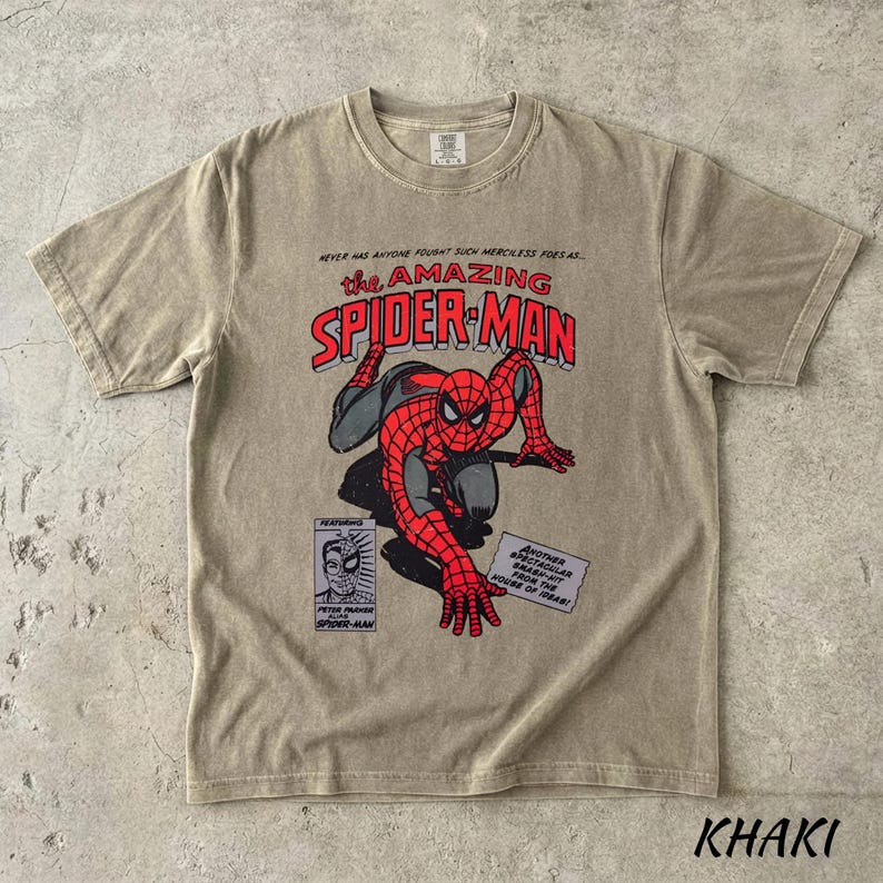 May include: Khaki-colored t-shirt featuring a vintage comic book graphic of Spider-Man. The design includes the text "The Amazing Spider-Man" in red and black, along with comic book panels. The shirt is a crew neck and has short sleeves.