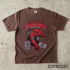 May include: Espresso-colored t-shirt featuring a vintage comic book graphic of Spider-Man. The design includes the text "The Amazing Spider-Man" in red and white, with Spider-Man in his classic red and blue suit. The shirt has a crew neck and short sleeves.