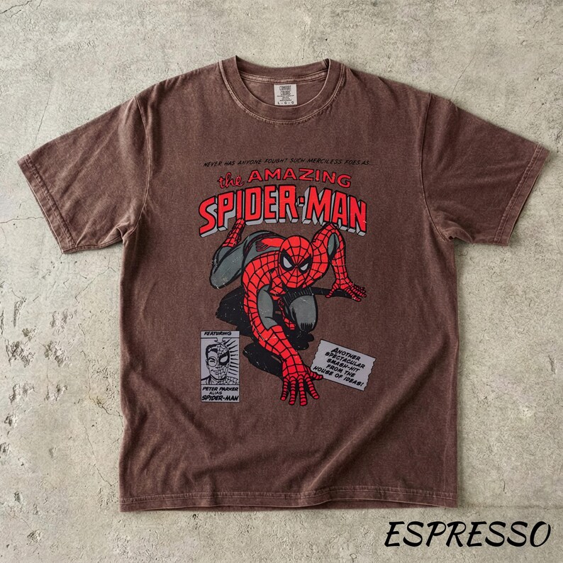 May include: Espresso-colored t-shirt featuring a vintage comic book graphic of Spider-Man. The design includes the text "The Amazing Spider-Man" in red and white, with Spider-Man in his classic red and blue suit. The shirt has a crew neck and short sleeves.