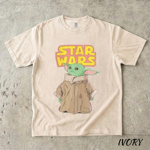 May include: Ivory-colored t-shirt featuring a graphic of Grogu from Star Wars. The design includes the Star Wars logo in yellow and pink. Grogu is depicted in a brown coat with a green face and large ears.