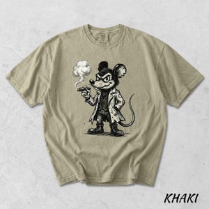 May include: Khaki-colored t-shirt featuring a black and white cartoon illustration of a mouse smoking a cigar. The mouse is dressed in a coat and boots. The word "KHAKI" is printed in black at the bottom of the shirt.