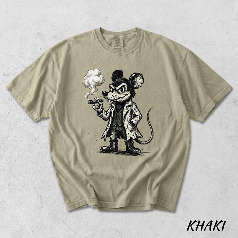 May include: Khaki-colored t-shirt featuring a black and white cartoon illustration of a mouse smoking a cigar. The mouse is dressed in a coat and boots. The word "KHAKI" is printed in black at the bottom of the shirt.