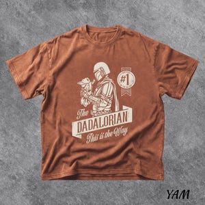 Pu&ograve; includere: Una maglietta marrone con una grafica di The Mandalorian che tiene Grogu, con la scritta "The Dadalorian This is the Way". Il design include un distintivo "Certified #1 Dad". La maglietta ha un aspetto vintage e usurato.