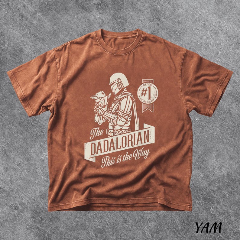 Pu&ograve; includere: Una maglietta marrone con una grafica di The Mandalorian che tiene Grogu, con la scritta "The Dadalorian This is the Way". Il design include un distintivo "Certified #1 Dad". La maglietta ha un aspetto vintage e usurato.