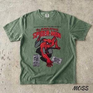 May include: A moss green t-shirt featuring a graphic of Spider-Man in red and black, with the text "the AMAZING SPIDER-MAN" in red. Additional text includes "NEVER HAS ANYONE FOUGHT SUCH MERCILESS FOES AS..." and "MOSS".
