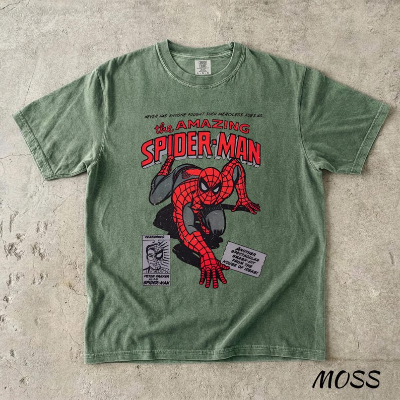 May include: A moss green t-shirt featuring a graphic of Spider-Man in red and black, with the text "the AMAZING SPIDER-MAN" in red. Additional text includes "NEVER HAS ANYONE FOUGHT SUCH MERCILESS FOES AS..." and "MOSS".