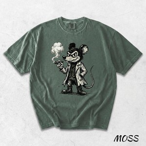 May include: Moss green t-shirt featuring a cartoon mouse smoking a cigar. The mouse is dressed in a coat and boots, with a stern expression. The shirt has a crew neck and short sleeves. The word "MOSS" is printed at the bottom right.