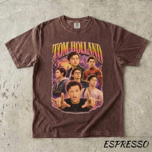 May include: Espresso-colored t-shirt featuring a graphic of Tom Holland in various poses, with the text "TOM HOLLAND" in a retro font. The shirt has a crew neck and short sleeves, and the word "ESPRESSO" is printed at the bottom.