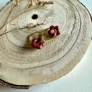 Op de afbeelding: Een paar gouden oorringen met bordeauxrode bloemvormige bedels. De oorbellen worden getoond op een rustieke houten schijf, met gedroogde bloemen. Een uniek accessoire.