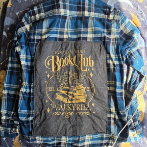 May include: A blue and white plaid flannel shirt featuring a large, rectangular dark gray fabric patch sewn onto the back. The patch displays gold text reading House of Wind Book Club, Est. 21, and Valkyrie reading room, with a stack of books illustration.