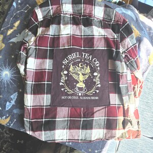 May include: A long-sleeve button-down flannel shirt in a burgundy, black, and white plaid pattern. A large dark brown square patch is sewn on the back featuring a yellow graphic of a tea cup, mountains, and text reading Suriel Tea Co.