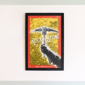 May include: A framed poster with a red background and a yellow pattern of rocks. A pickaxe is held by a gloved hand. The text "Rock and Stone" is written in a stylized font. The text "Property of Deep Rock Galactic" is written at the bottom of the poster.