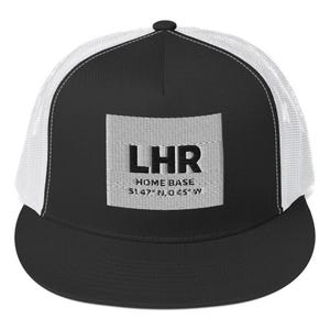 Pode incluir: Boné de caminhoneiro preto e branco com aba curva. O painel frontal apresenta um patch retangular com as letras "LHR", as palavras "HOME BASE" e coordenadas. O boné tem uma parte traseira de malha e um fecho de pressão ajustável.