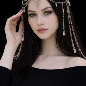 May include: Silver-toned headpiece with multiple chains and dangling teardrop-shaped charms. The headpiece is worn over dark hair, with the chains cascading down. The woman is wearing a black off-the-shoulder top.