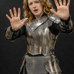 May include: A suit of polished steel plate armor featuring a breastplate, pauldrons, and vambraces worn over a black long-sleeved shirt. The armor includes a chainmail coif and a chainmail skirt with metal rings, secured by dark leather straps.