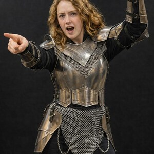 May include: A suit of polished steel plate armor featuring a breastplate, pauldrons, and vambraces worn over a black long-sleeve shirt. The armor includes a chainmail hauberk, tassets, and leather straps with metal buckles, providing full torso coverage.