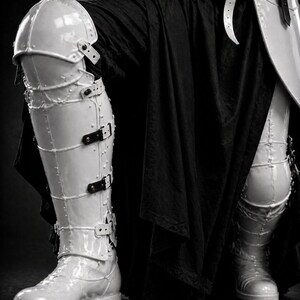 May include: Close-up of white, segmented armor plating, including leg and shoulder protection. The armor is secured with black straps and buckles. A black fabric drapes behind the armor, creating a contrast. The image is in black and white.