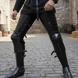 Puede incluir: Armadura de pierna medieval negra con placas articuladas y correas de cuero. La armadura cubre los muslos, las rodillas y las espinillas, y se usa sobre pantalones y botas negros. La armadura est&aacute; dise&ntilde;ada para la protecci&oacute;n y una est&eacute;tica hist&oacute;rica.