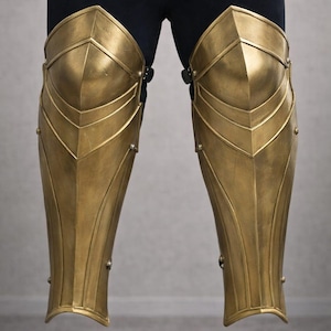 Puede incluir: Armadura de pierna dorada, con un diseño segmentado con detalles remachados. La armadura parece ser de metal y se usa sobre leggings negros. Se ven las rodilleras y las espinilleras.