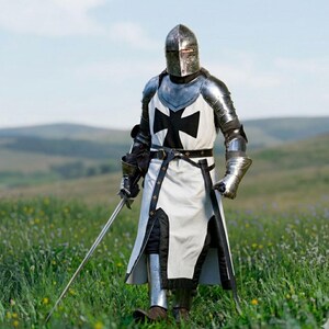 Puede incluir: Un caballero medieval con armadura completa est&aacute; en un campo de flores silvestres. La armadura del caballero es plateada y negra, con una t&uacute;nica blanca con una gran cruz negra. El caballero sostiene una espada larga y un casco cubre su rostro.