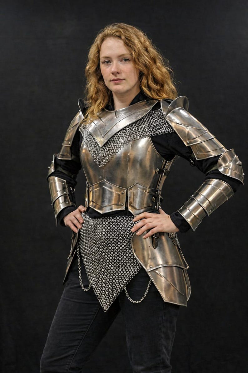 May include: A silver-colored medieval armor set, including a chest plate, shoulder guards, and arm protection. The armor is detailed with chainmail and is worn over a black shirt and pants. The person is standing with their hands on their hips.