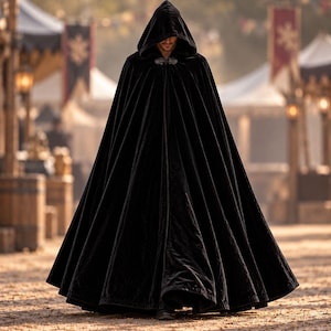 May include: A floor-length black velvet cloak featuring a deep hood and a decorative silver-toned metal clasp at the neckline. The garment has a full, flowing silhouette with vertical folds that extend from the shoulders to the hemline.