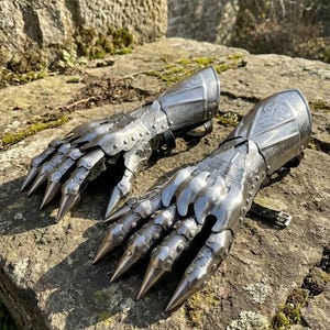 May include: A pair of silver-colored metal gauntlets with articulated fingers and long, pointed claws. The armor features decorative rivets and engraved patterns on the upper arm sections. The gauntlets are displayed on a rough, stone surface.