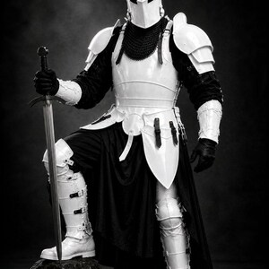 May include: A full suit of white and black medieval armor, including a helmet, chest plate, and leg guards. The knight holds a sword and stands on a rock. The armor is highly detailed and appears to be made of metal.