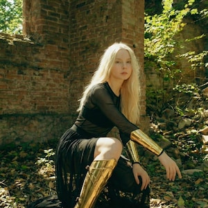 May include: A person wearing a black dress and gold armor, including arm and leg guards. The person is kneeling in a forest setting with a brick wall in the background. The armor has a metallic sheen.