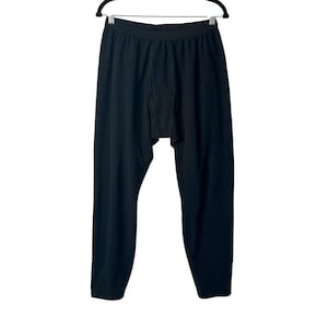 May include: A pair of solid black thermal pants featuring a textured fabric, an elasticized waistband, and a reinforced front fly panel. The garment is displayed hanging from a black plastic hanger, showing full-length legs with a straight cut.