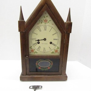 May include: A wooden Seth Thomas clock with a cream-colored face and Roman numerals. The clock has a dark brown frame with two pointed spires and floral accents. A key rests below the clock.