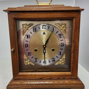 May include: A wooden mantel clock with a gold-colored face and black numerals. The clock has a rectangular shape with a handle on top and decorative gold accents. The clock is made of wood and has a glass front.