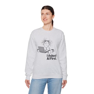 May include: Light grey sweatshirt featuring a cartoon graphic of a person working on a laptop, with the text "I Asked AI First." The graphic includes a laptop and two coffee mugs. The sweatshirt is worn by a person wearing blue jeans.