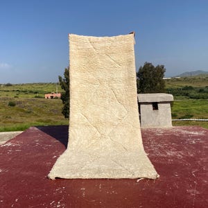 May include: A beige, rectangular rug with a geometric pattern is displayed outdoors. The rug is made of a thick, textured material and is draped over a red surface. The background features a clear blue sky, green fields, and a small building.