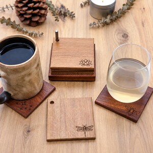 May include: A set of square wooden coasters featuring laser-engraved designs including a lotus flower, a dragonfly, a bee, and starbursts. The coasters are dark brown with a smooth finish, some stacked in a holder with a metal pin, and others placed individually under a ceramic mug and a clear glass.